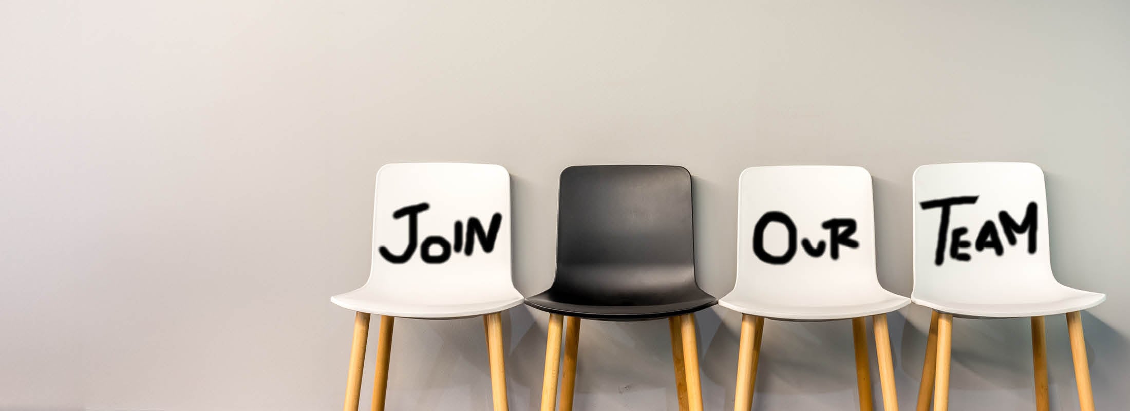 Vier Stühle stehen nebeneinander vor einer hellgrauen Wand, auf drei von ihnen steht der Schriftzug „Join Our Team“.
