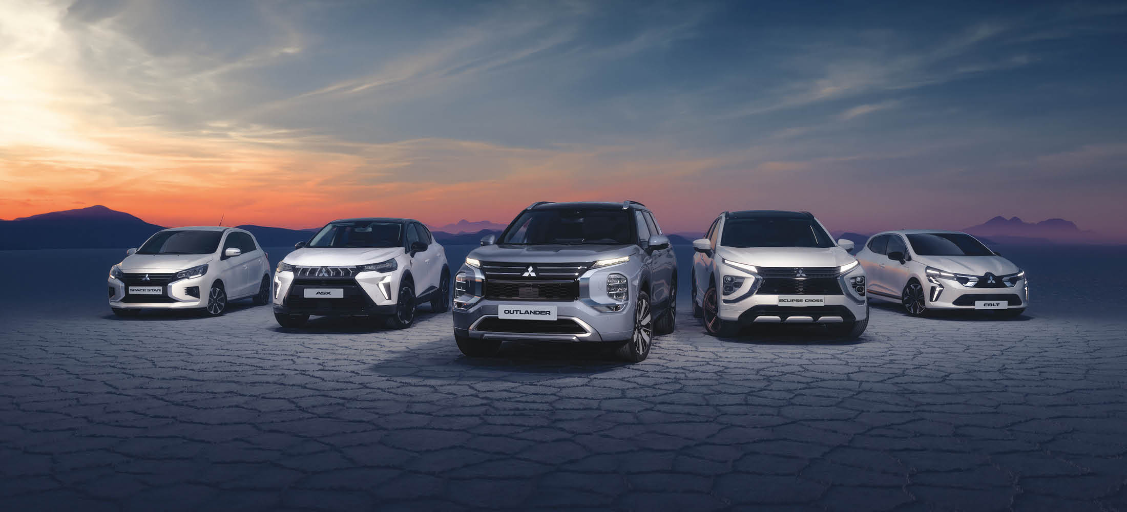 Fünf Mitsubishi-Modelle stehen nebeneinander in einer weiten Landschaft bei Sonnenuntergang.