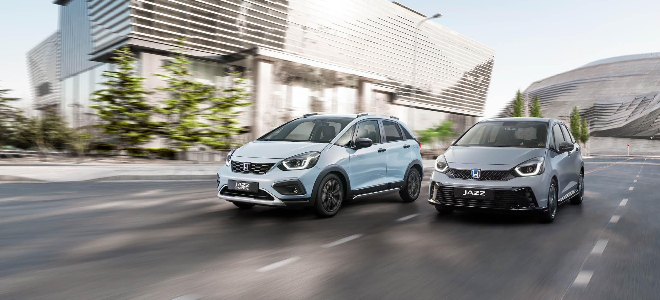 Zwei Honda Jazz-Modelle fahren nebeneinander auf einer modernen, städtischen Straße mit futuristisch wirkenden Gebäuden im Hintergrund.