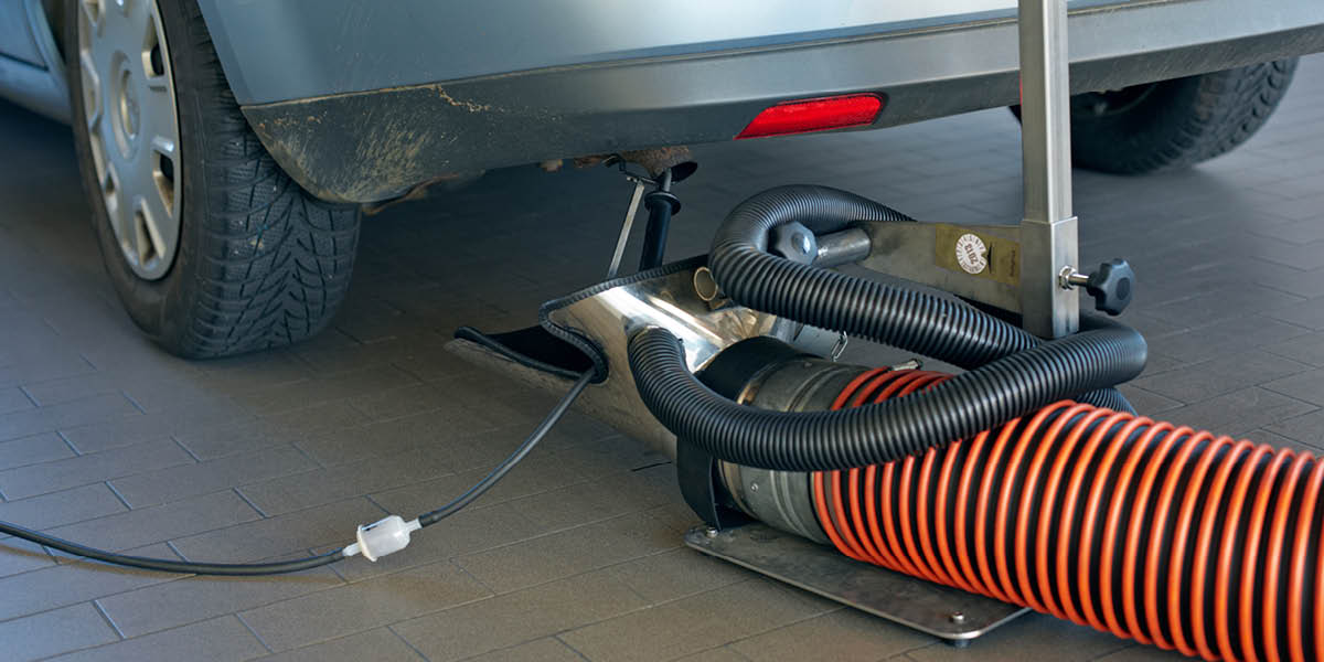 Ein Auto steht mit dem Heck im Bildvordergrund. Am Auspuff ist ein Abgasmessgerät mit einem orange-schwarzen Schlauch angeschlossen.